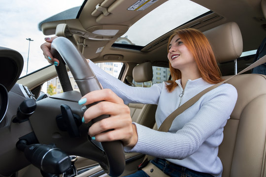 Wide angle view of young redhead woman driver fastened by seatbelt driving a car smiling happily.